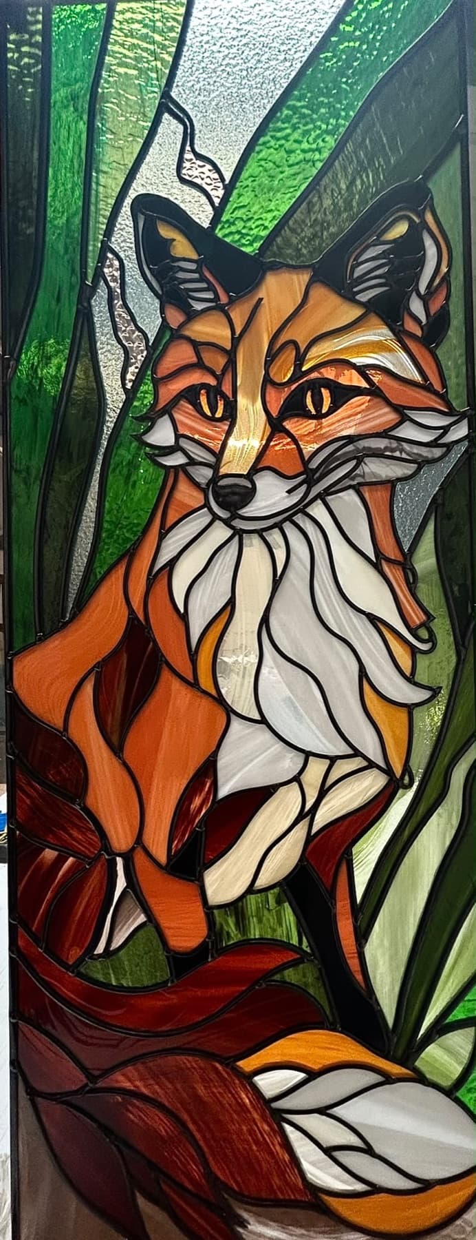 A fox sitting among green reeds, in orange, cream and green glass