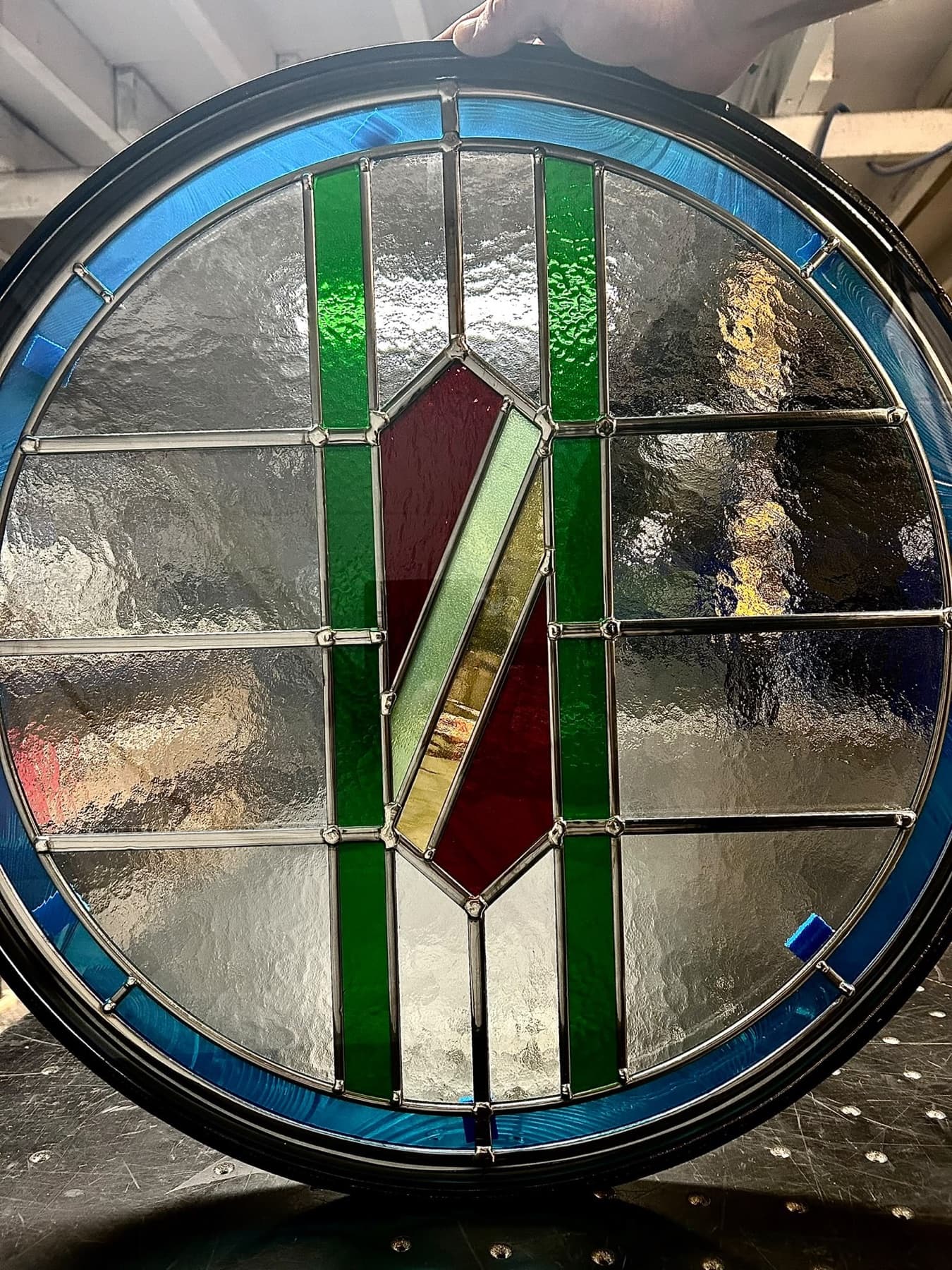 A round leaded unit in clear textured glass with green bars and a red and amber centre, held up in the workshop